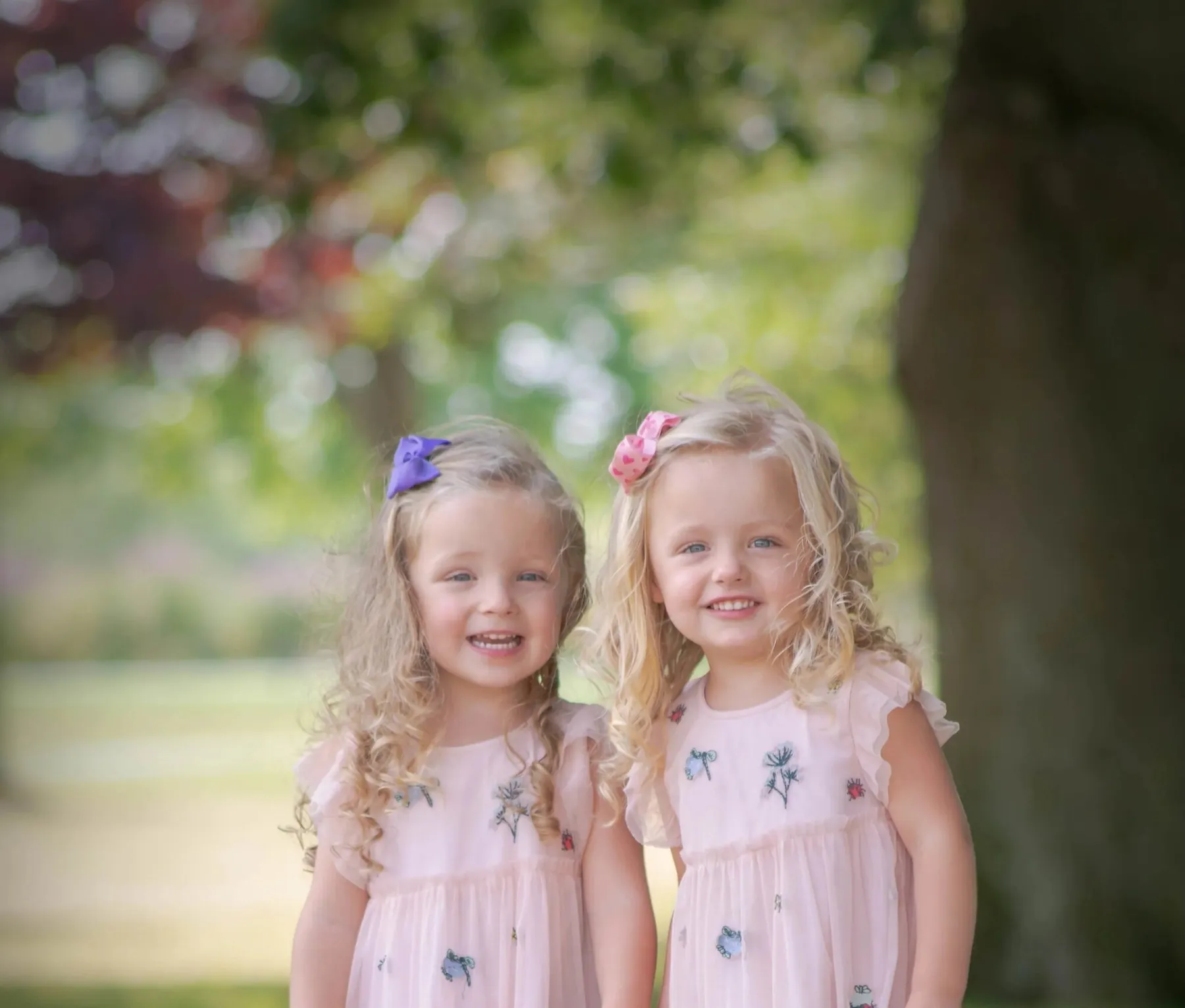 Two young sisters in matching pale pink dresses with flowers in their hair, standing under a tree in summer