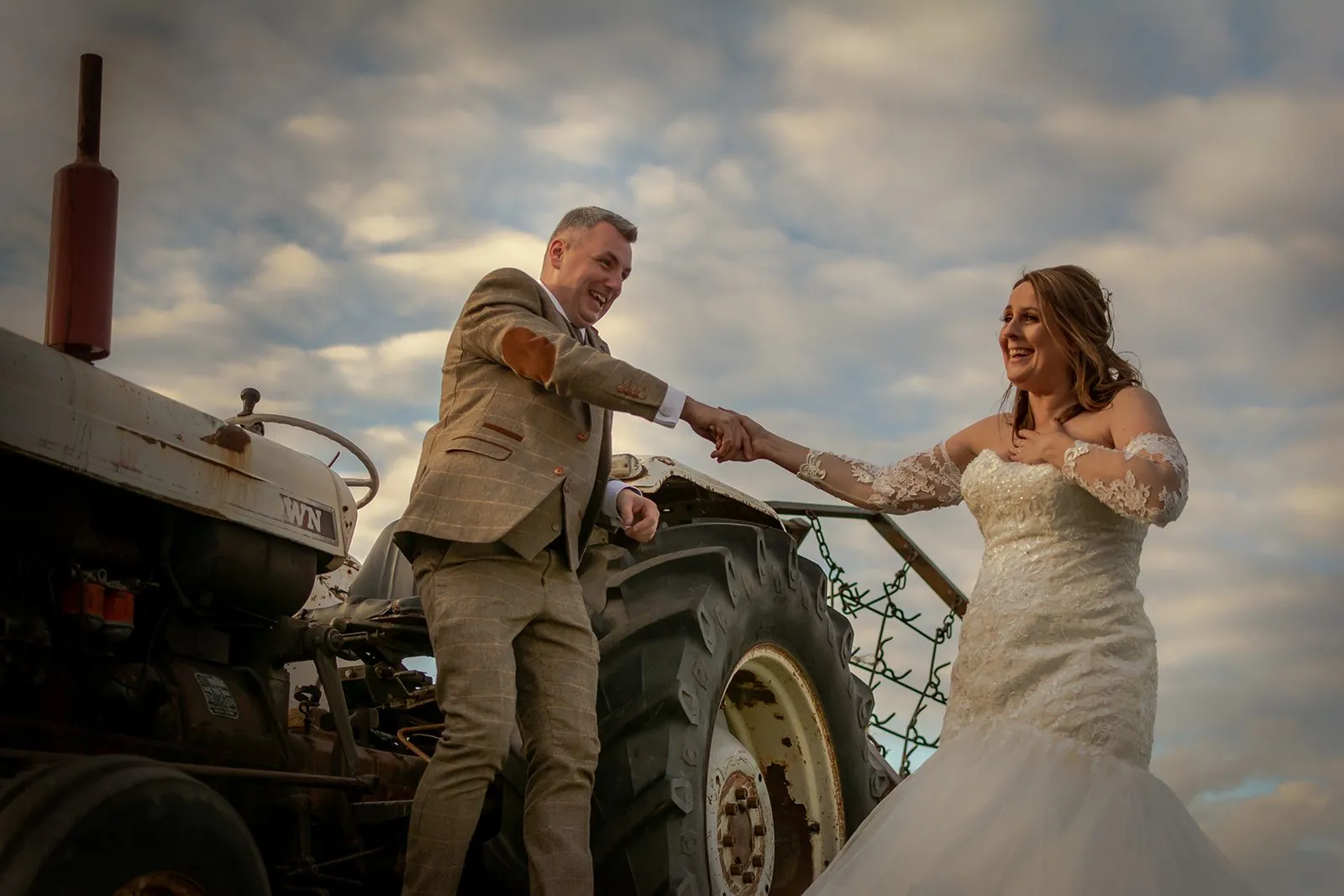 Groom sitting on an old grey farm tractor reaching down to hold the bride's hand, cloud piled up behind them